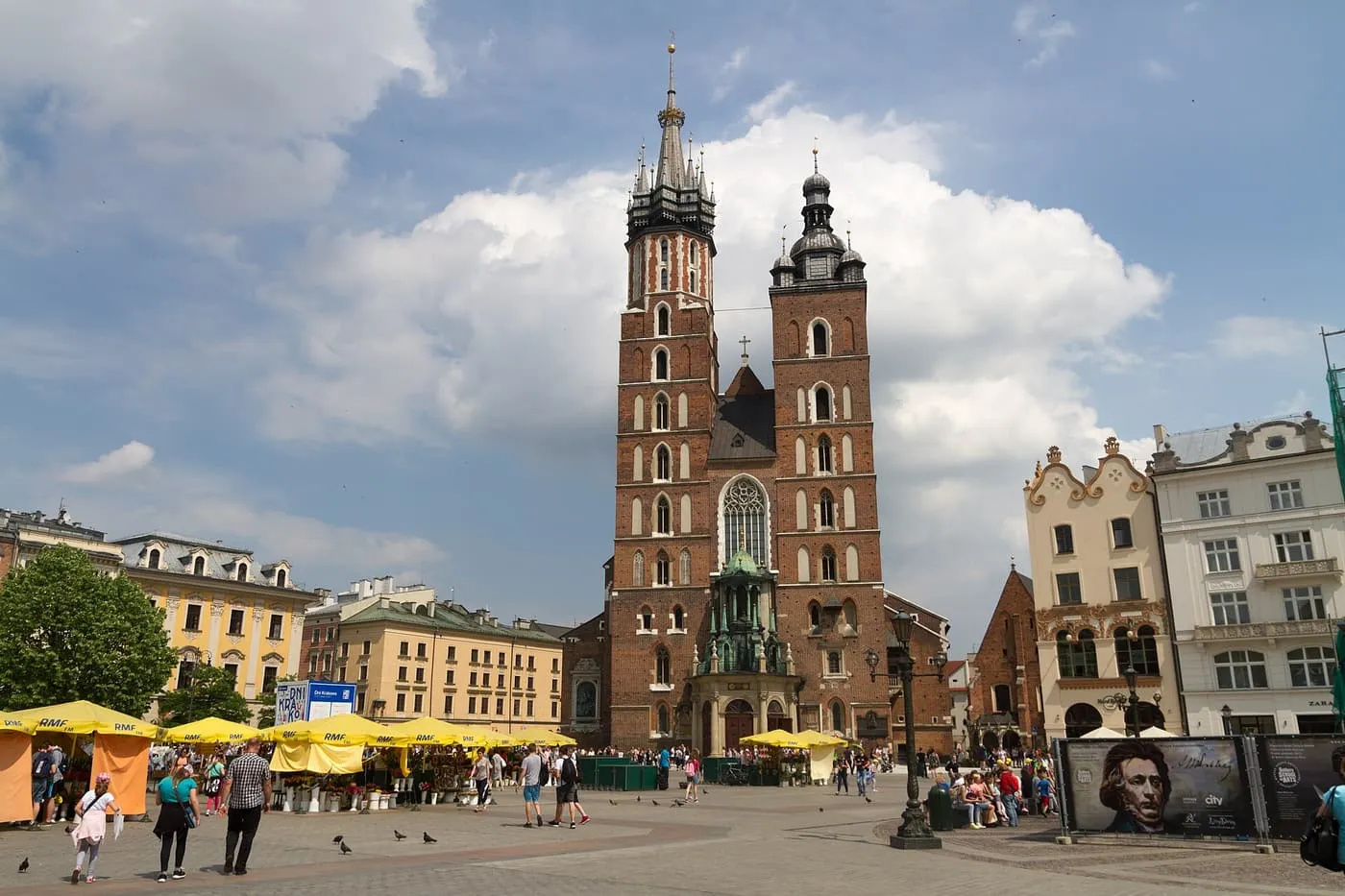 The two brick towers of St Mary's Basilica, one taller with a spire, above the market stalls and people on the Main Market Square