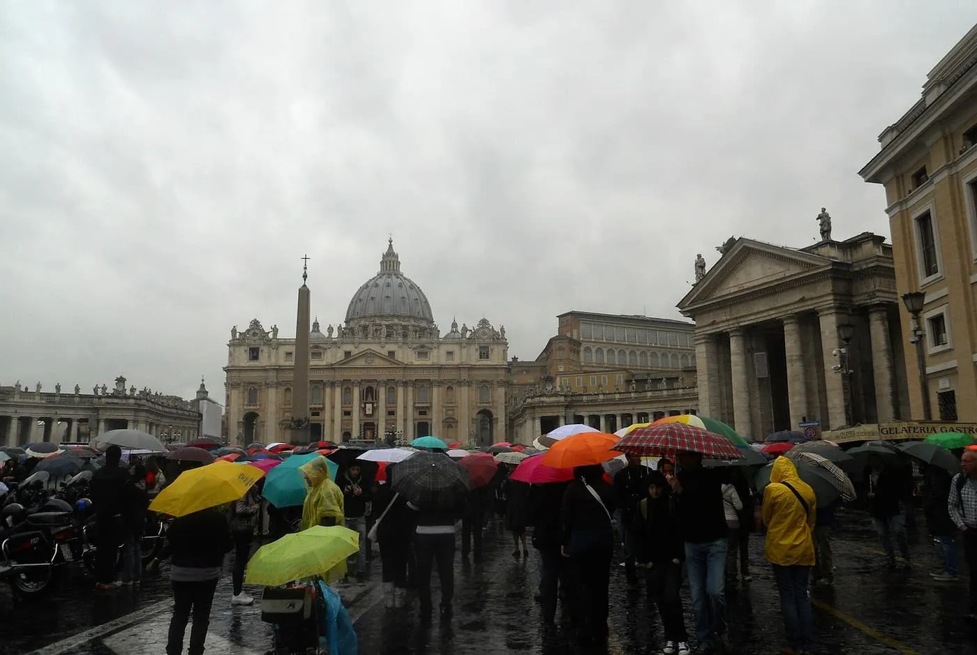 Crowds under colourful umbrellas on wet cobbles in St Peter's Square, with the basilica dome under a grey sky