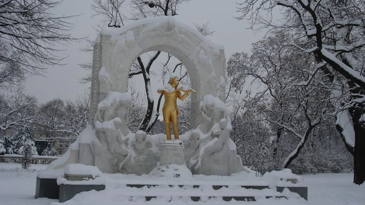 The gilded statue of Johann Strauss playing the violin under a snow-covered marble arch in the Stadtpark, with snowy trees behind
