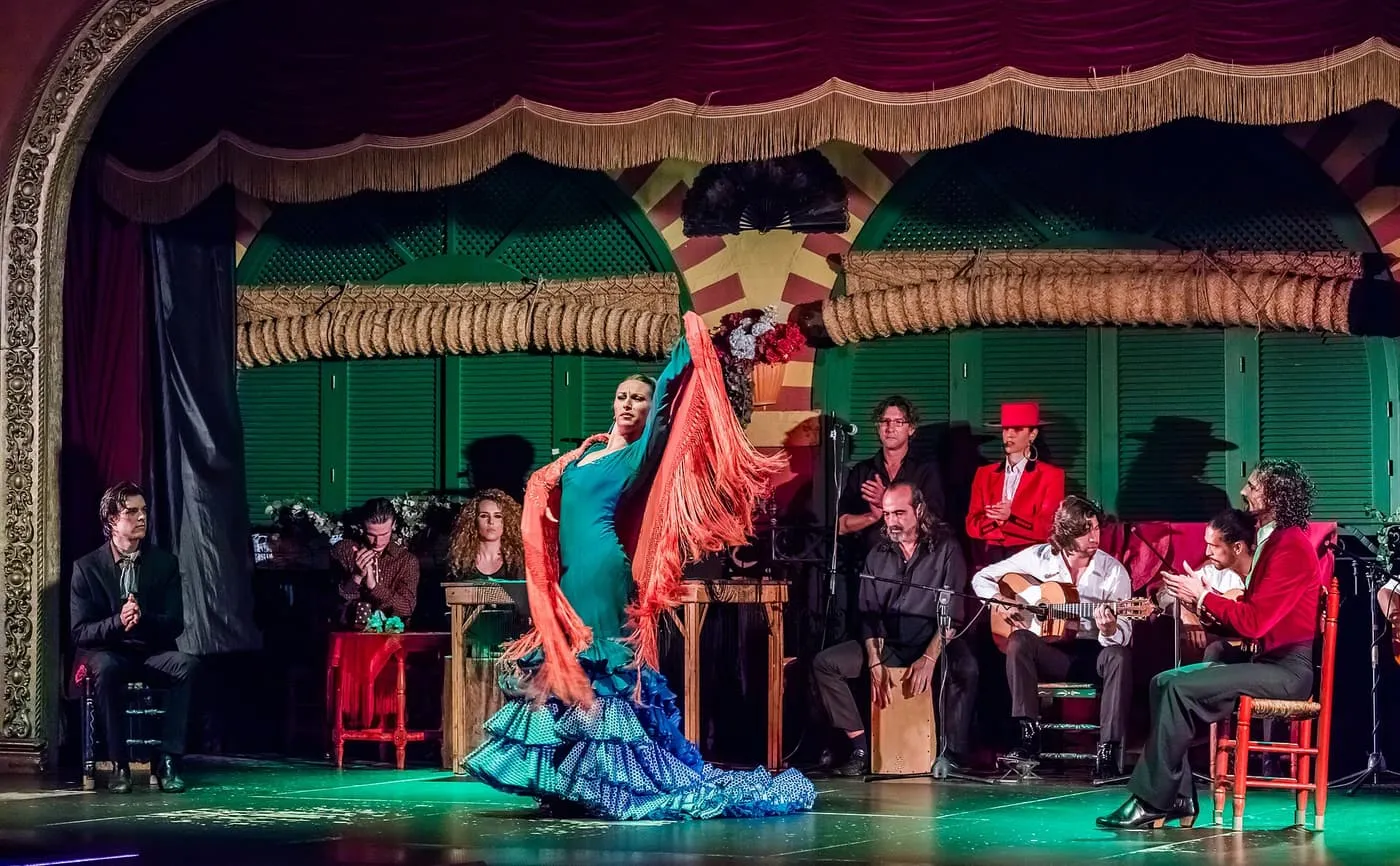 A dancer in a blue dress and orange shawl on a stage with green shutters, with guitarists, a cajón player and singers seated behind her