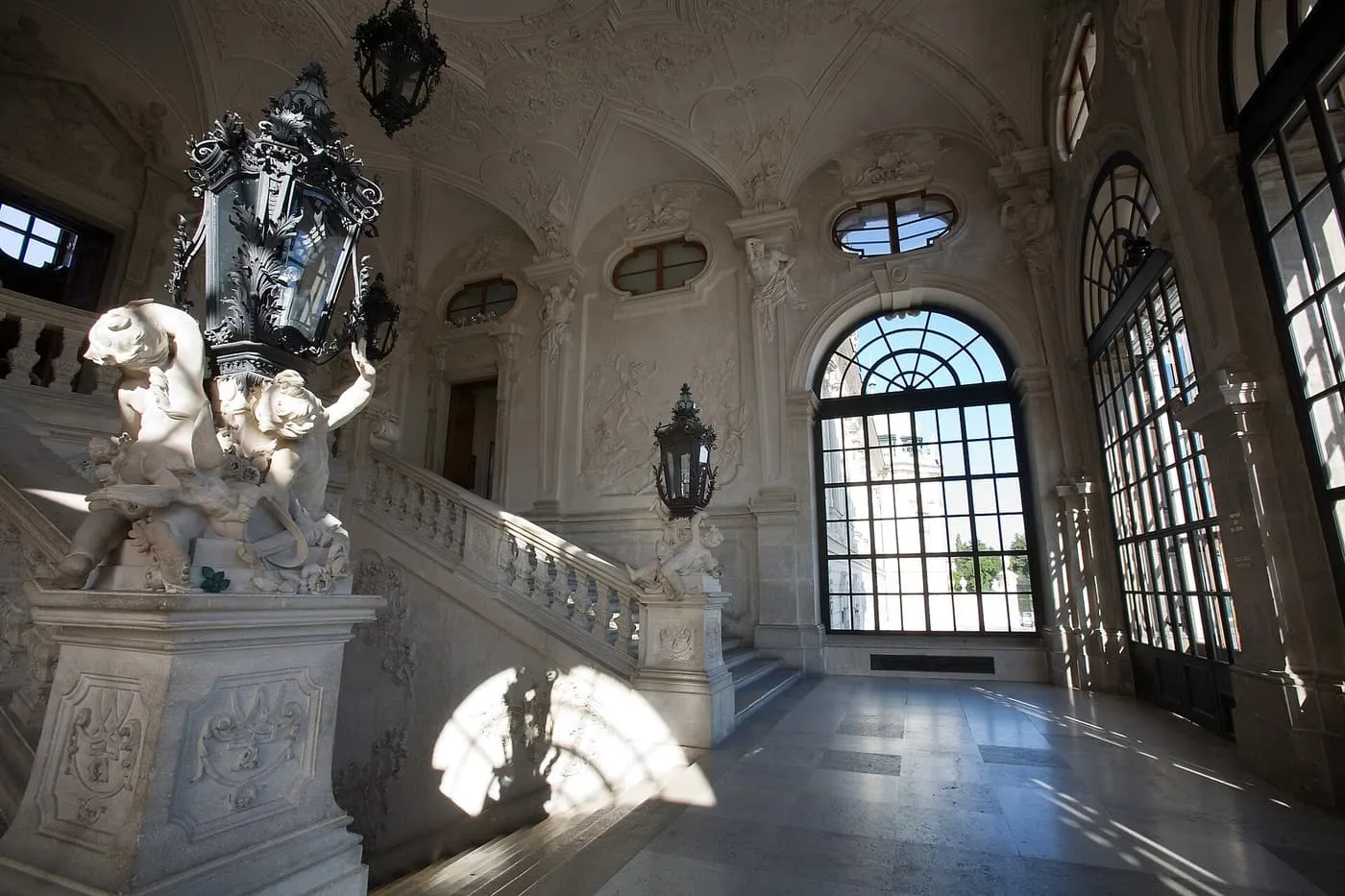 A stone staircase hall with a white balustrade, putti carrying a wrought-iron lantern and tall arched windows