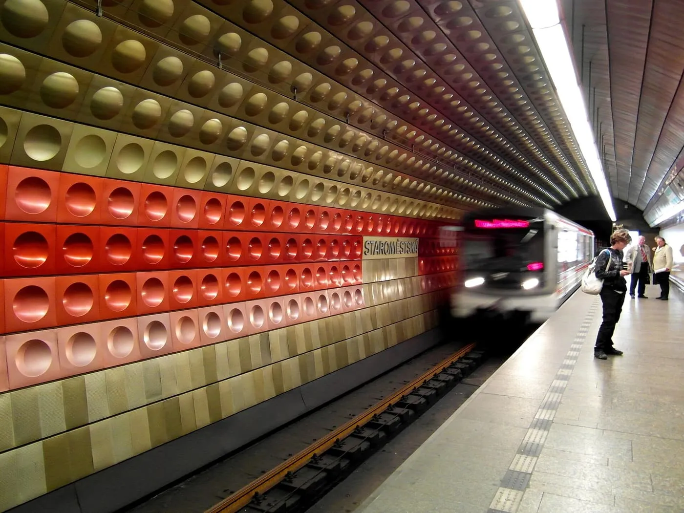 A metro train arriving at Staroměstská station, whose walls are clad in red and gold dished panels, with a few passengers waiting on the platform