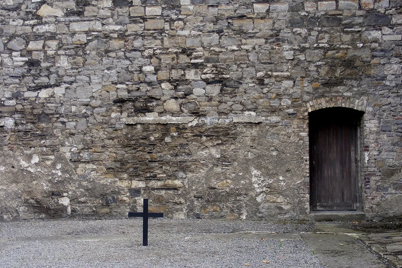 A rough stone wall in the Stonebreakers' Yard at Kilmainham Gaol, with a wooden door and a black cross standing in the gravel