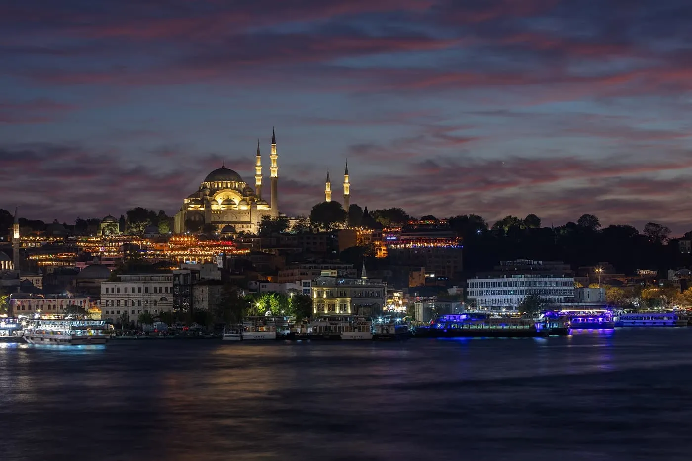 The lit domes and minarets of the Süleymaniye Mosque on its hill at dusk, with boats on the Golden Horn below
