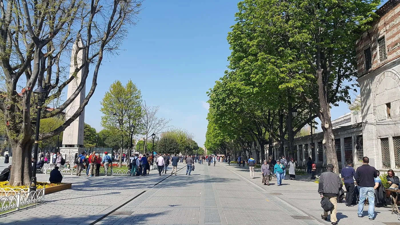 Sultanahmet Square on a sunny day, a wide paved space between rows of plane trees with an obelisk in the distance and visitors walking