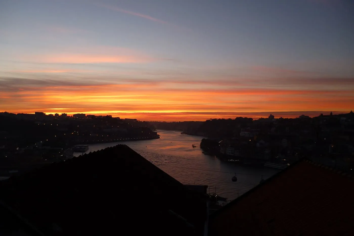 An orange and pink sunset over the Douro, with the river running between dark hills and two rooftops in the foreground