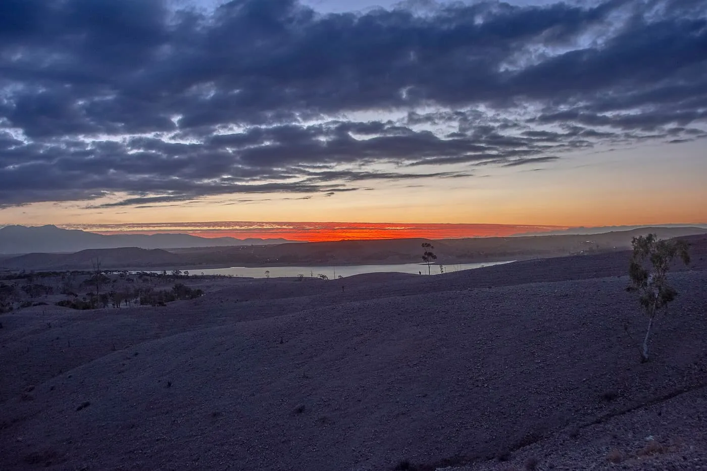 An orange band of sunset under dark cloud over bare hills near Agafay, with a stretch of water below