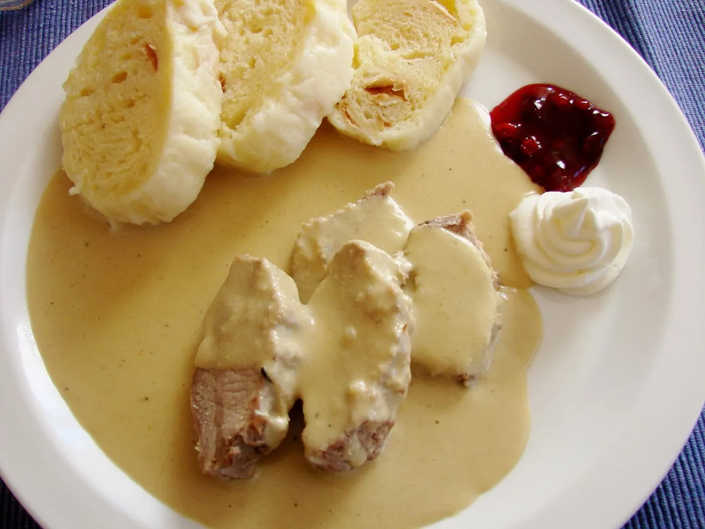 A plate of svíčková: slices of braised beef in a pale cream sauce with sliced bread dumplings, a spoon of cranberry jam and a swirl of whipped cream