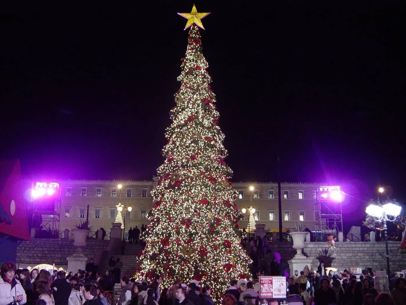 A tall Christmas tree covered in white lights and red decorations with a gold star, standing in front of the Parliament building on Syntagma Square at night