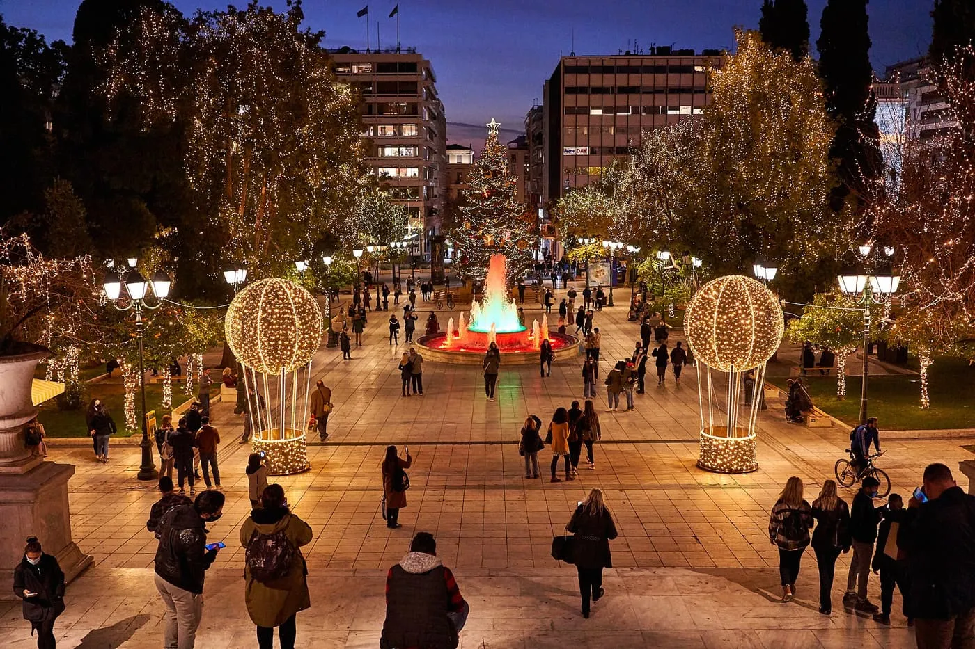 Syntagma Square at dusk seen from above, with trees wrapped in fairy lights, two lit balloon-shaped decorations, a fountain and a Christmas tree at the far end