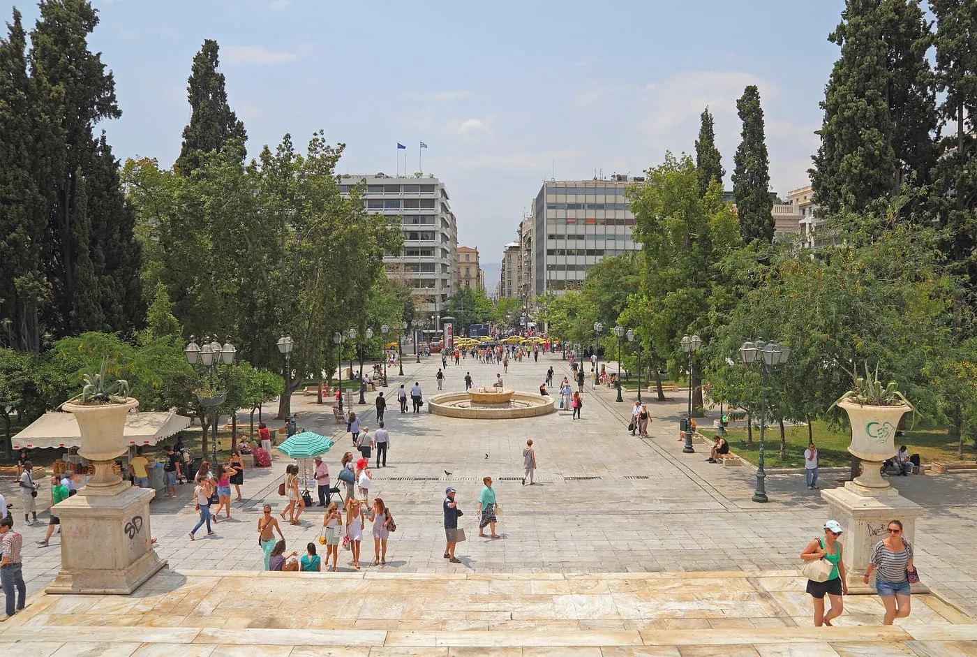 Syntagma Square seen from above, a wide paved square with a fountain in the middle, cypress trees on either side and people crossing in the sun