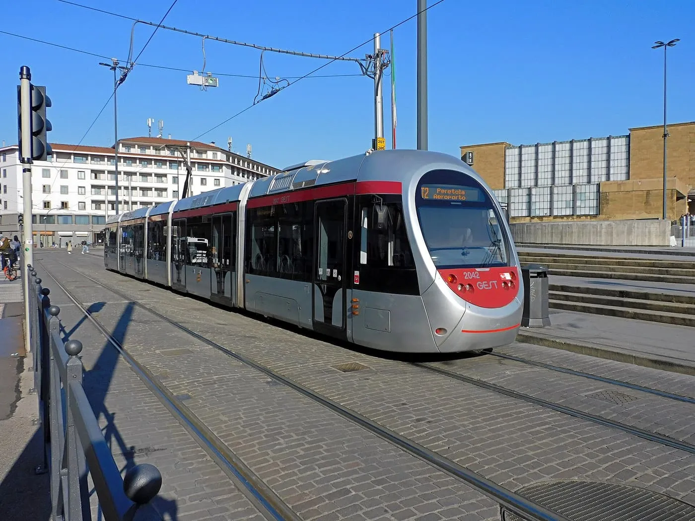 A silver and red T2 tram with Peretola Aeroporto on its destination board, crossing the paving of Piazza della Stazione