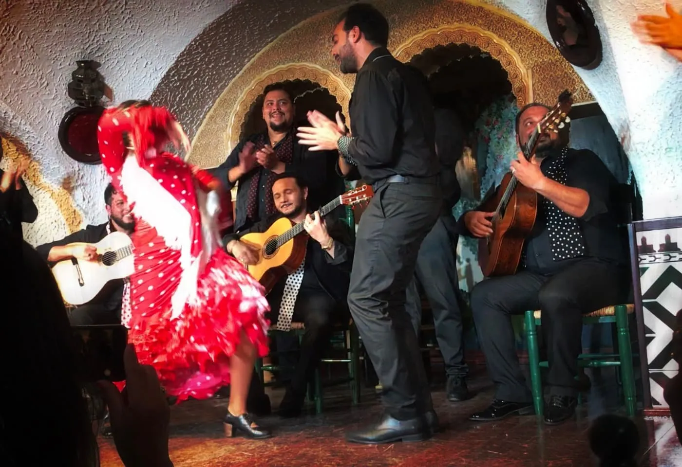 A flamenco dancer in a red polka-dot dress mid-turn beside two guitarists and a standing performer clapping, in front of Moorish-style arches
