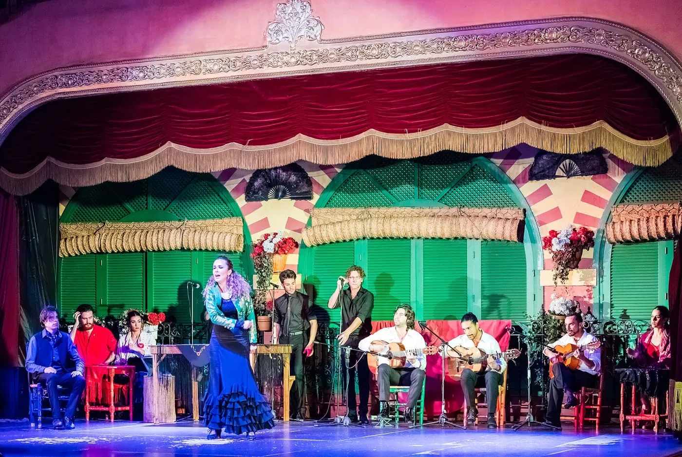 A singer in a blue dress in front of three guitarists on a tablao stage with a red velvet curtain and green shutters