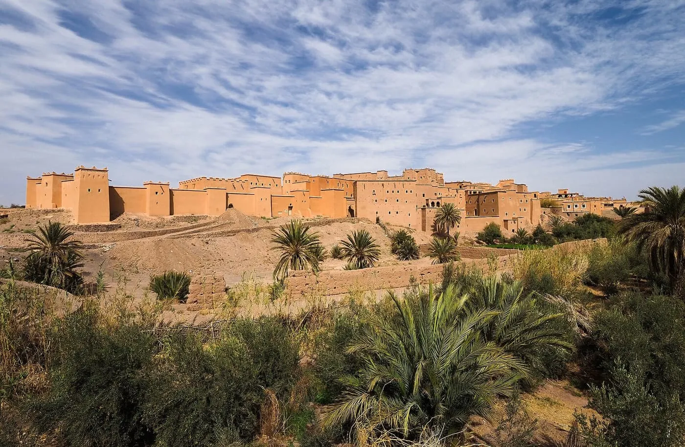 The long earthen walls and towers of Taourirt kasbah behind palms and scrub under a streaked sky