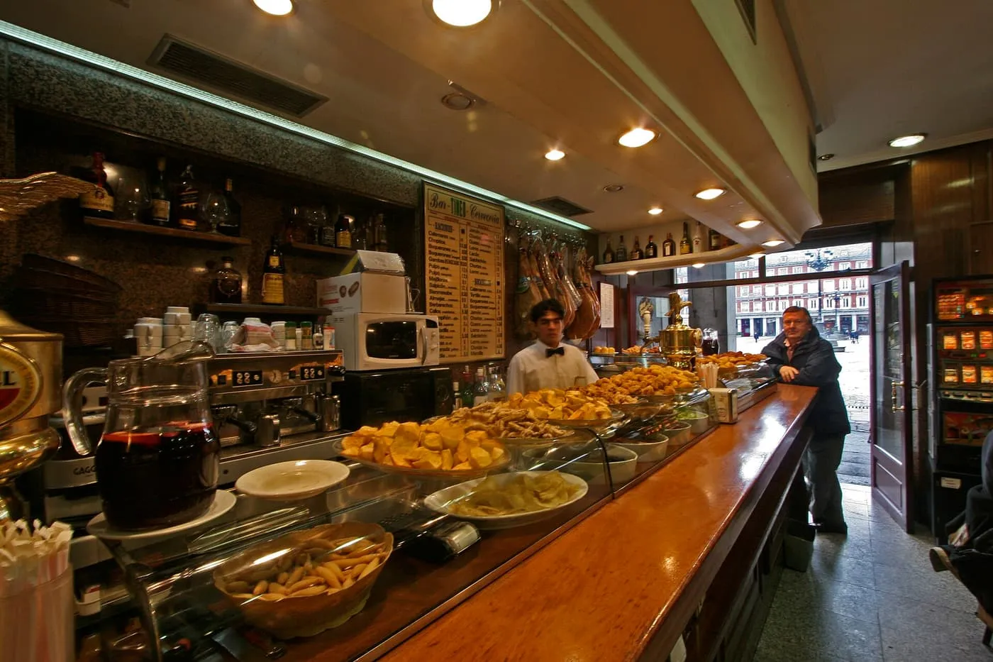 A tapas bar counter with dishes of fried potatoes, nuts and olives under glass, a jug of sangría, hams hanging behind and the Plaza Mayor visible through the open door