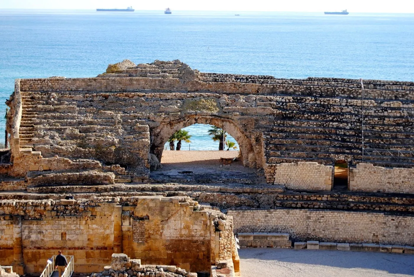 The stone tiers and central arch of Tarragona's Roman amphitheatre with the blue Mediterranean beyond and cargo ships on the horizon