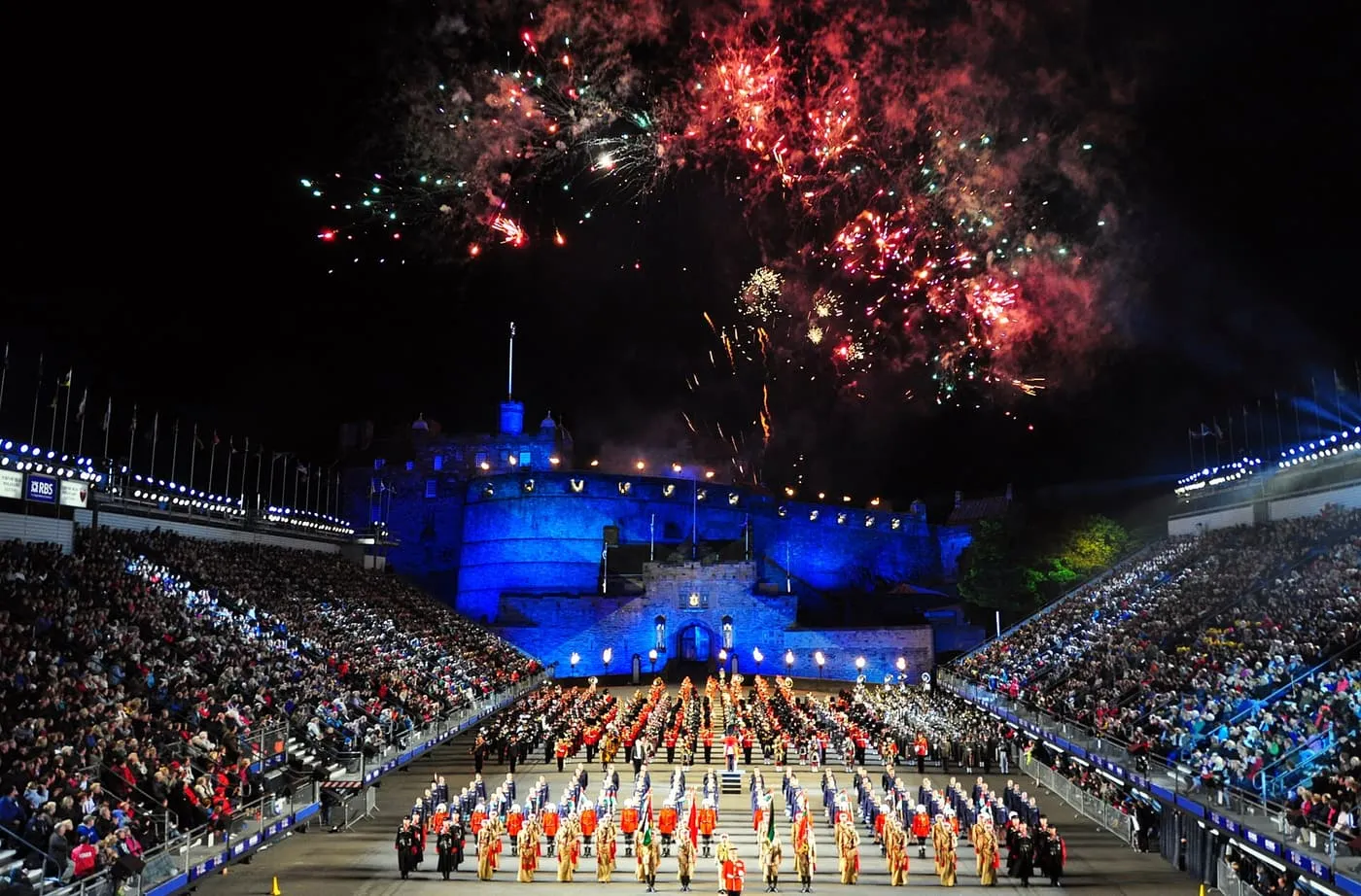 Massed bands on the Castle Esplanade in front of the floodlit blue castle gatehouse, with grandstands full on both sides and fireworks overhead