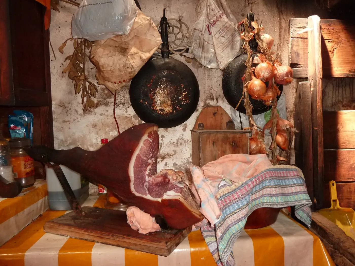 A cured ham on a wooden board beside a string of onions and old frying pans hung on the wall