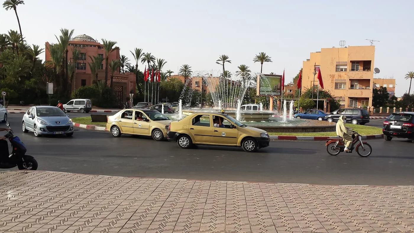 Two beige taxis passing a fountain on a palm-lined roundabout in Marrakech