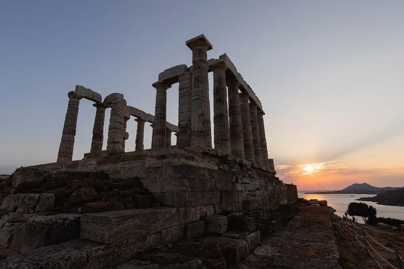 The columns of the Temple of Poseidon in silhouette at sunset, with the sun low over the sea and a headland beyond
