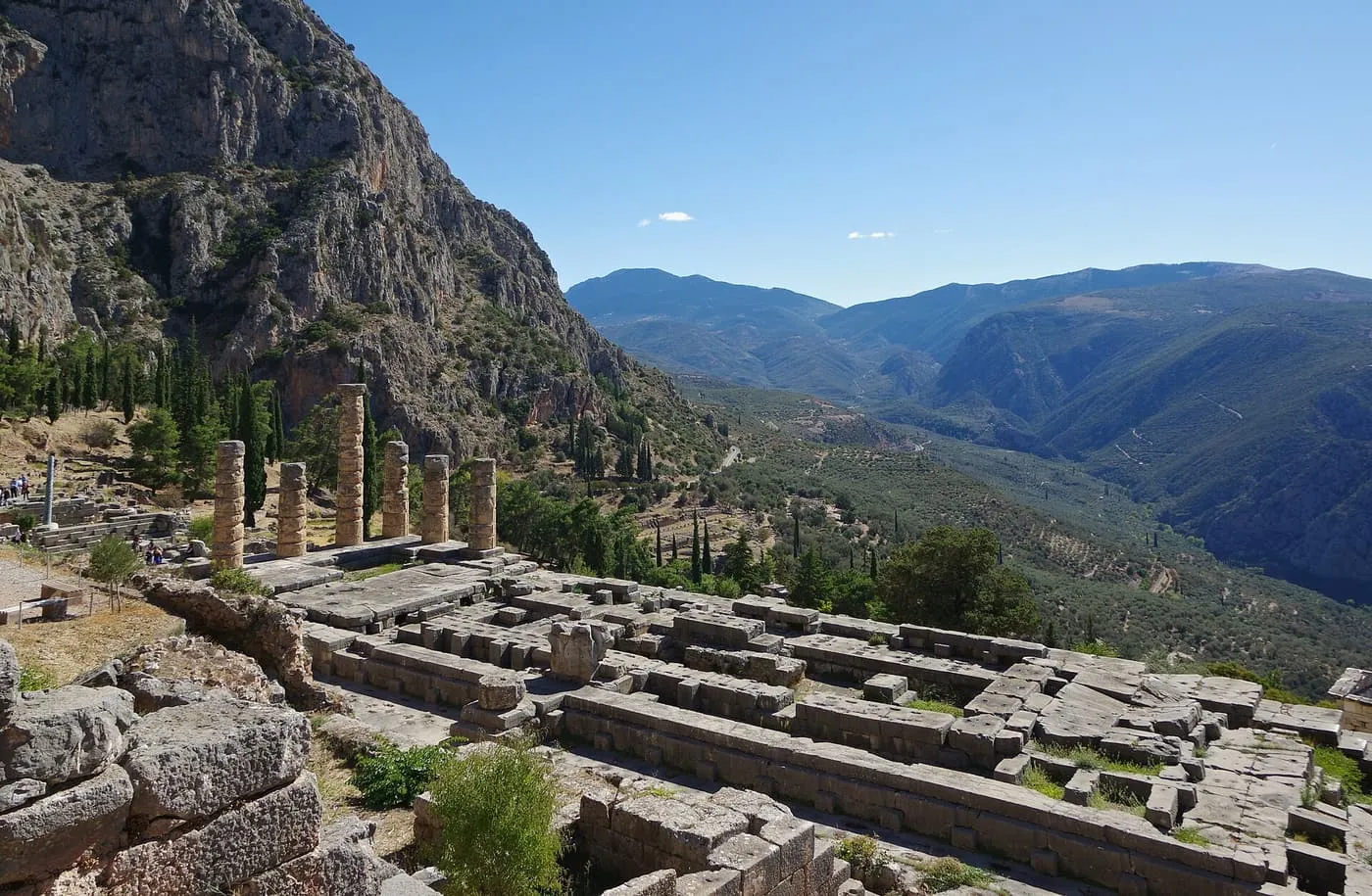 The standing columns of the Temple of Apollo at Delphi above the foundations of the temple, with a steep rock face and a valley beyond