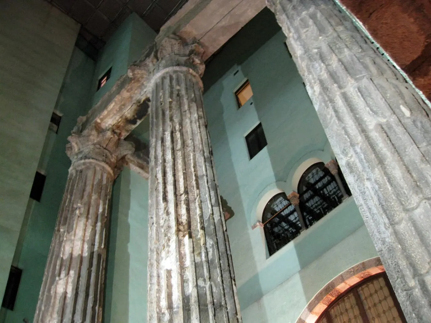 Looking up at three fluted Roman columns and a stretch of architrave inside a courtyard with pale green walls and arched windows