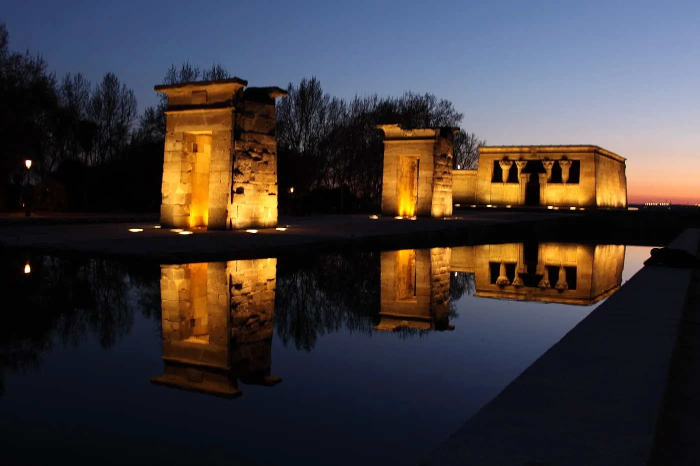 The Temple of Debod at dusk, its gateways and temple lit amber and reflected in the long pool in front of them