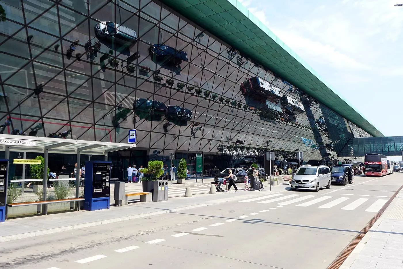 The glass front of the Kraków Airport terminal beside the road in front of it, with a bus stop and parked cars