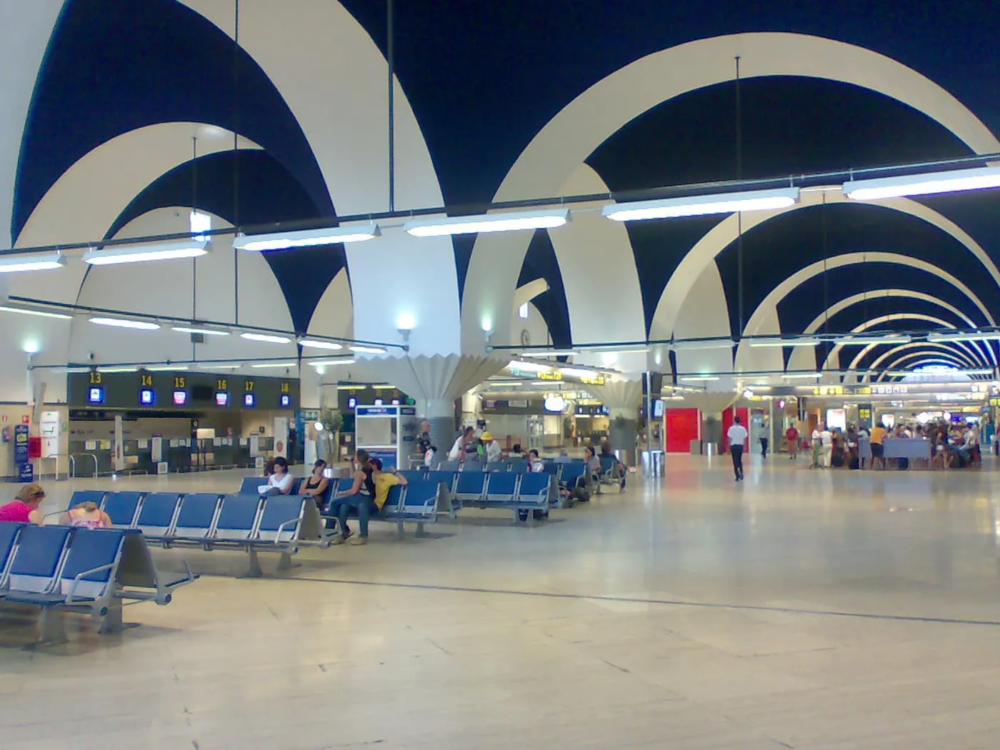 The hall of Seville Airport's terminal, with a ceiling of white arches over dark vaults, rows of blue seats and check-in desks