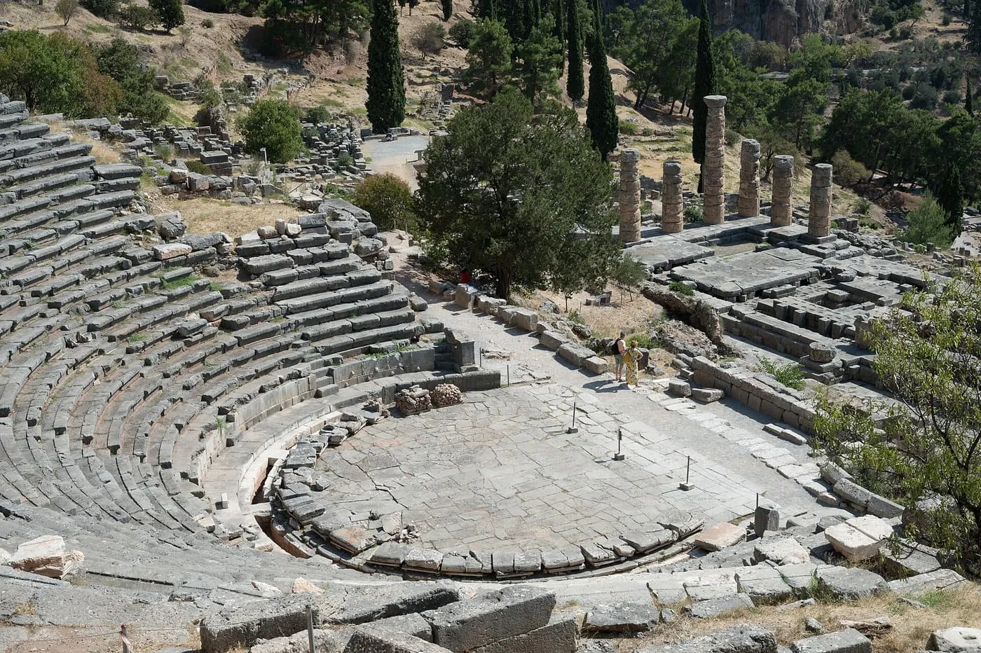 The stone seats and paved floor of the ancient theatre at Delphi seen from above, with the columns of the Temple of Apollo behind