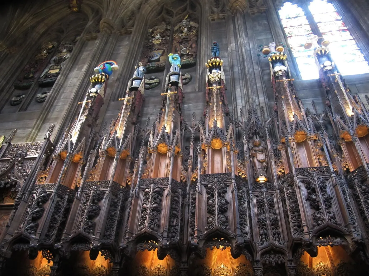 Carved oak stalls in the Thistle Chapel, with painted helms and crests on pinnacles above them and stained glass behind