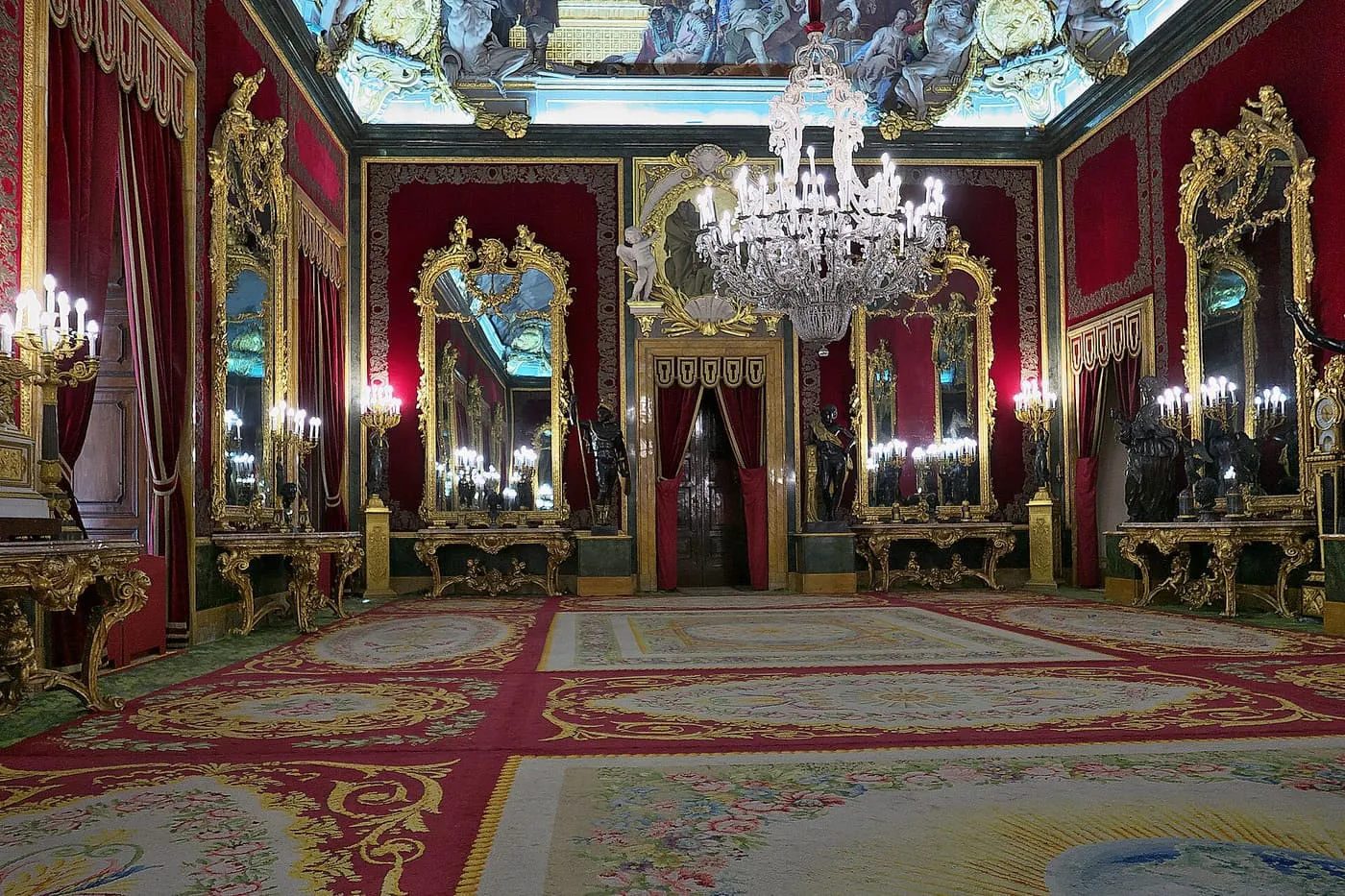 The Throne Room of the Royal Palace, with red velvet walls, gilded mirrors and consoles, a crystal chandelier and a painted ceiling above a patterned carpet