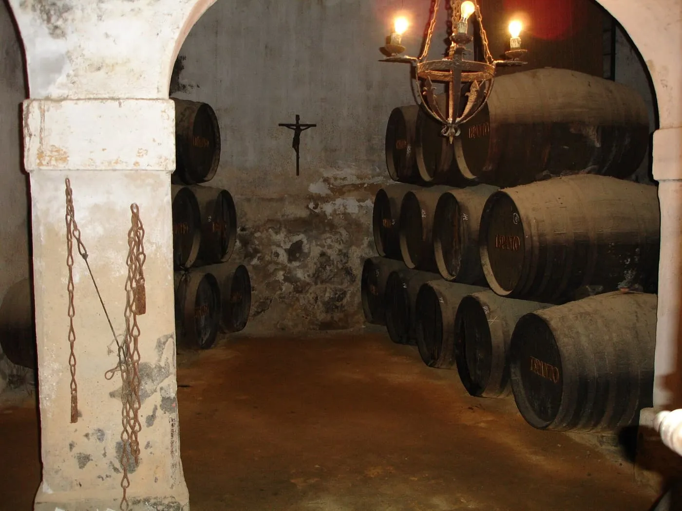 Sherry casks stacked in a whitewashed cellar under arches, with a candle-style lamp and a crucifix on the wall