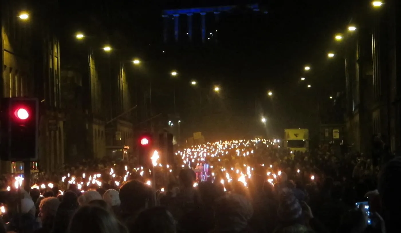 A dense crowd carrying flaming torches along a street at night, with traffic lights glowing red and a floodlit building above