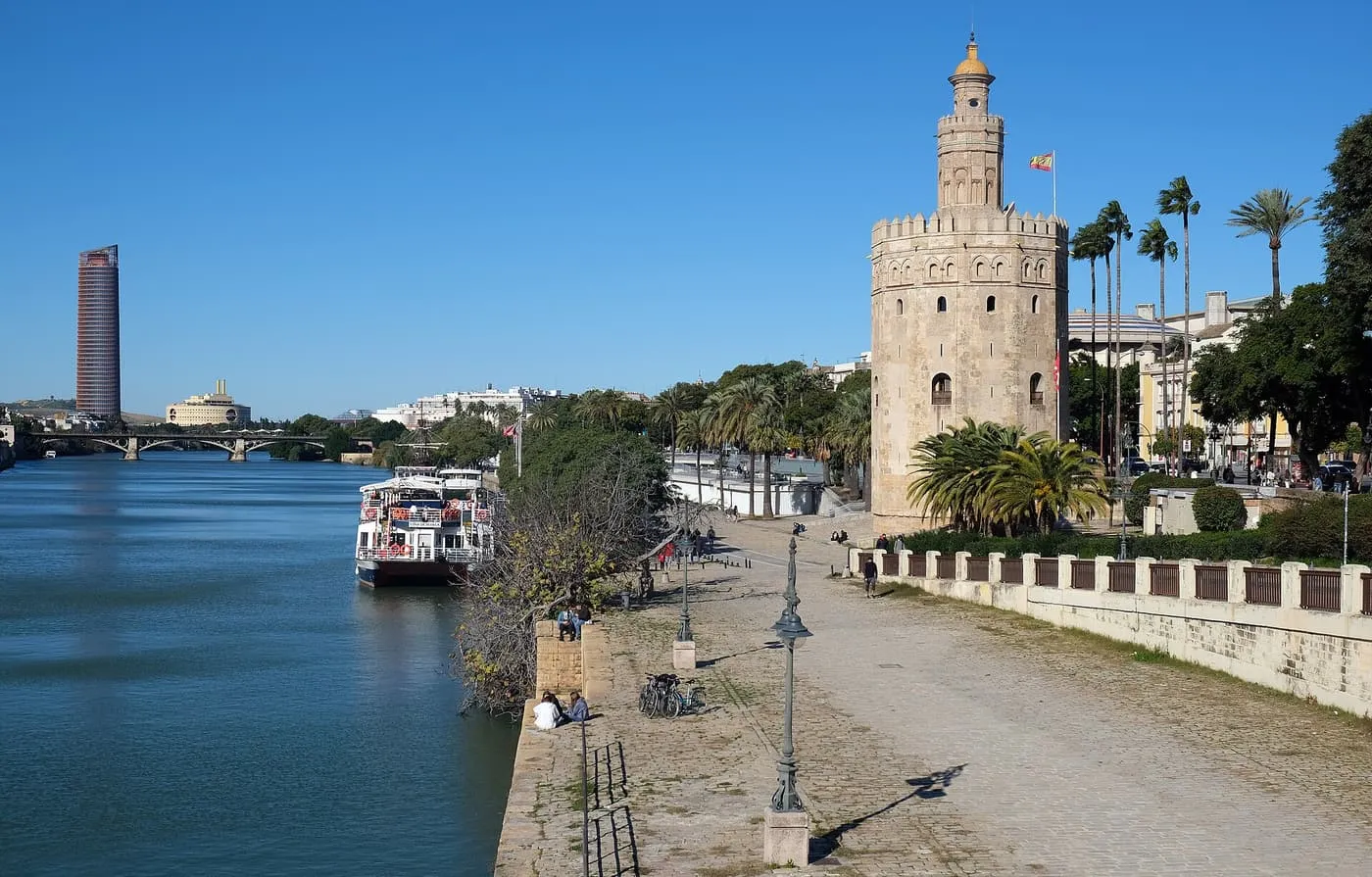 The Torre del Oro beside the Guadalquivir, with a river boat moored along the embankment and a tall tower in the distance under a blue sky