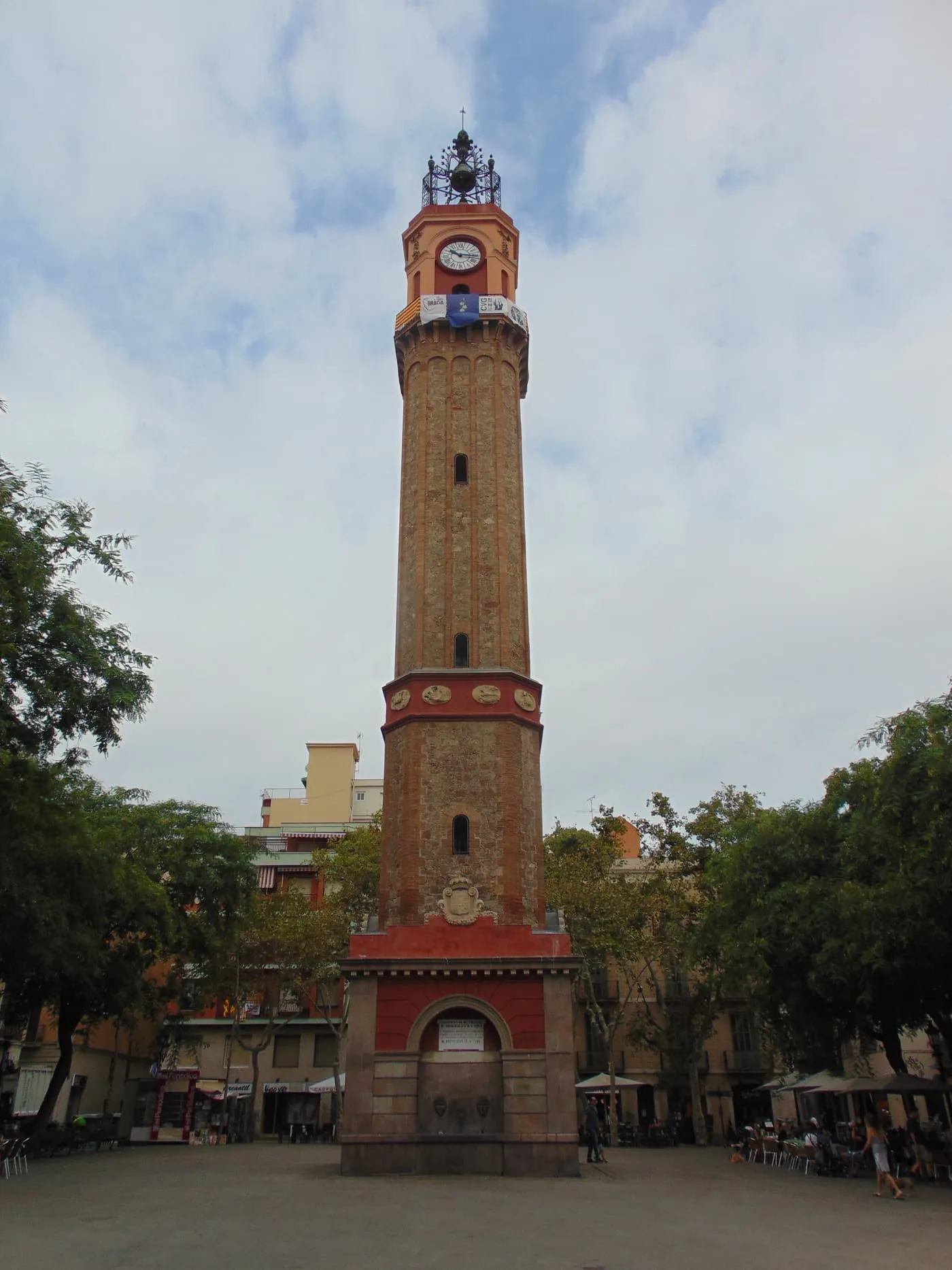 The brick clock tower in the middle of Plaça de la Vila de Gràcia, with a red base, café terraces and trees round the square