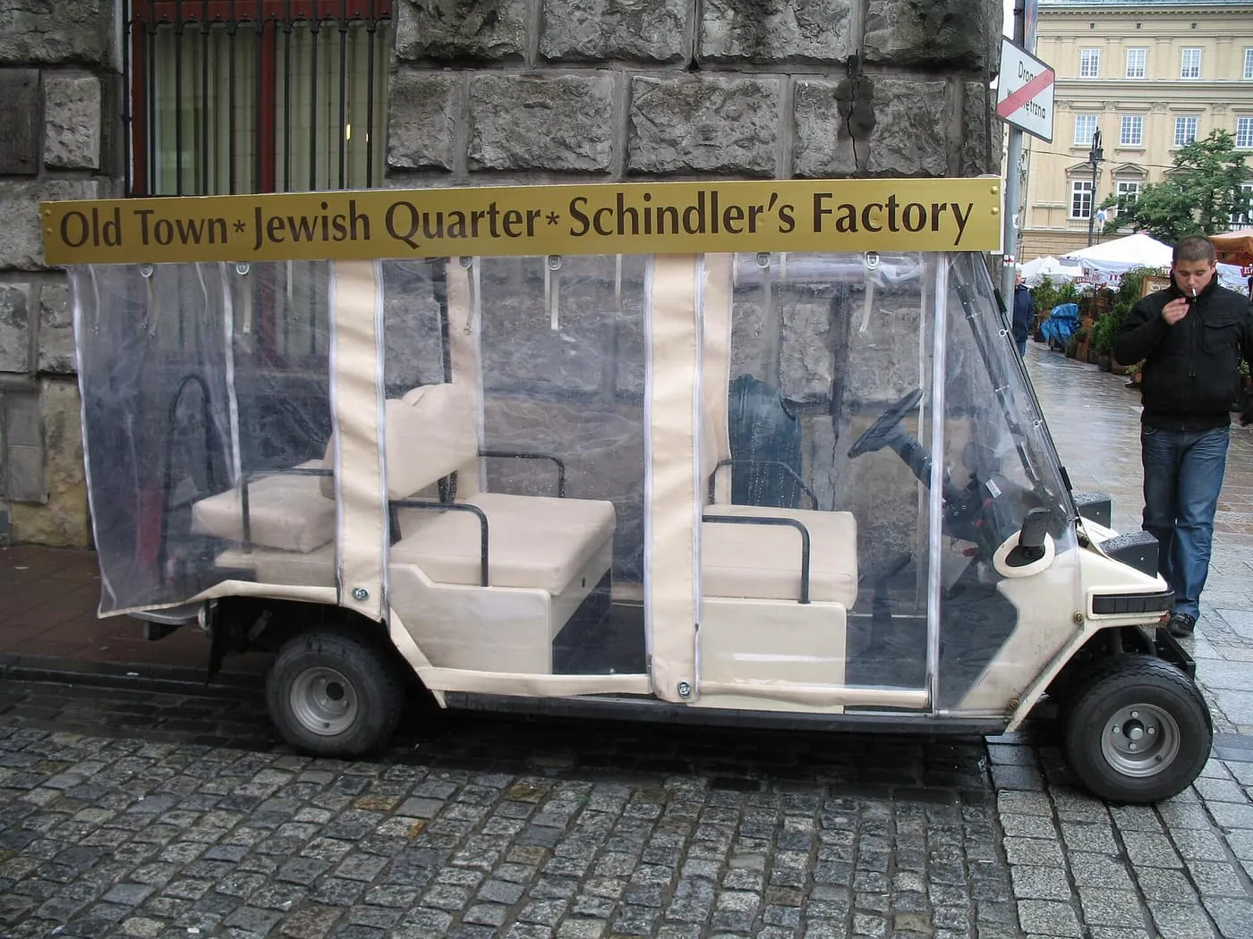 A white electric tourist cart with clear plastic side curtains and a gold roof sign reading Old Town, Jewish Quarter, Schindler's Factory, parked on cobbles