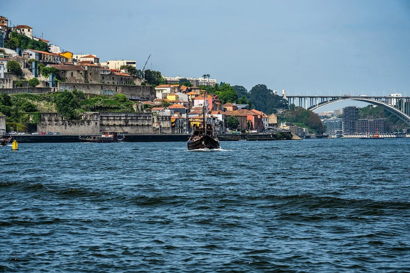 A dark wooden traditional boat under way on the blue-green Douro, with a hillside of old houses and the Arrábida bridge in the distance