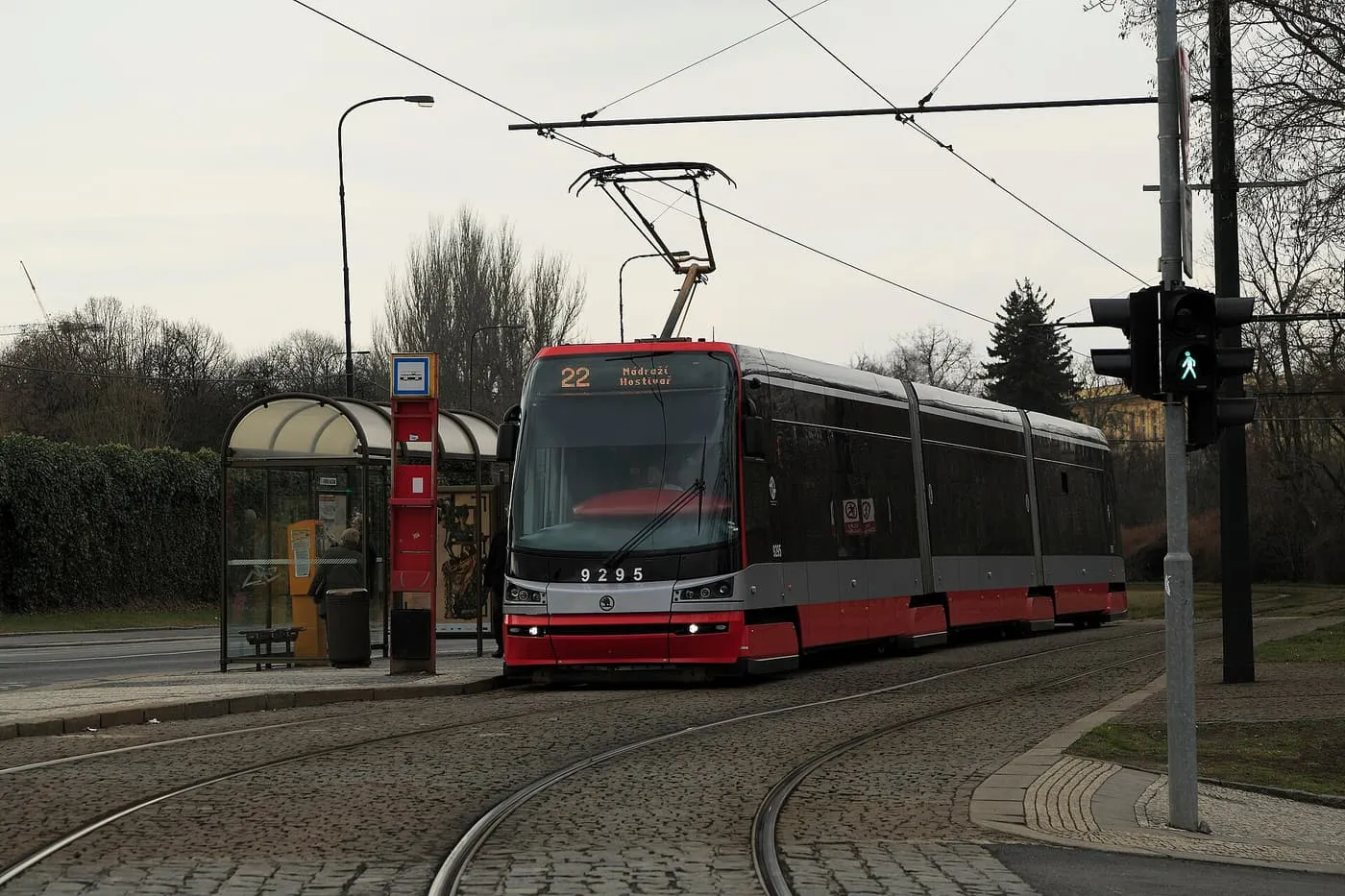 A red and silver Škoda 15T tram on line 22 standing at a tram stop with a glass shelter, beside a pedestrian crossing and cobbled track