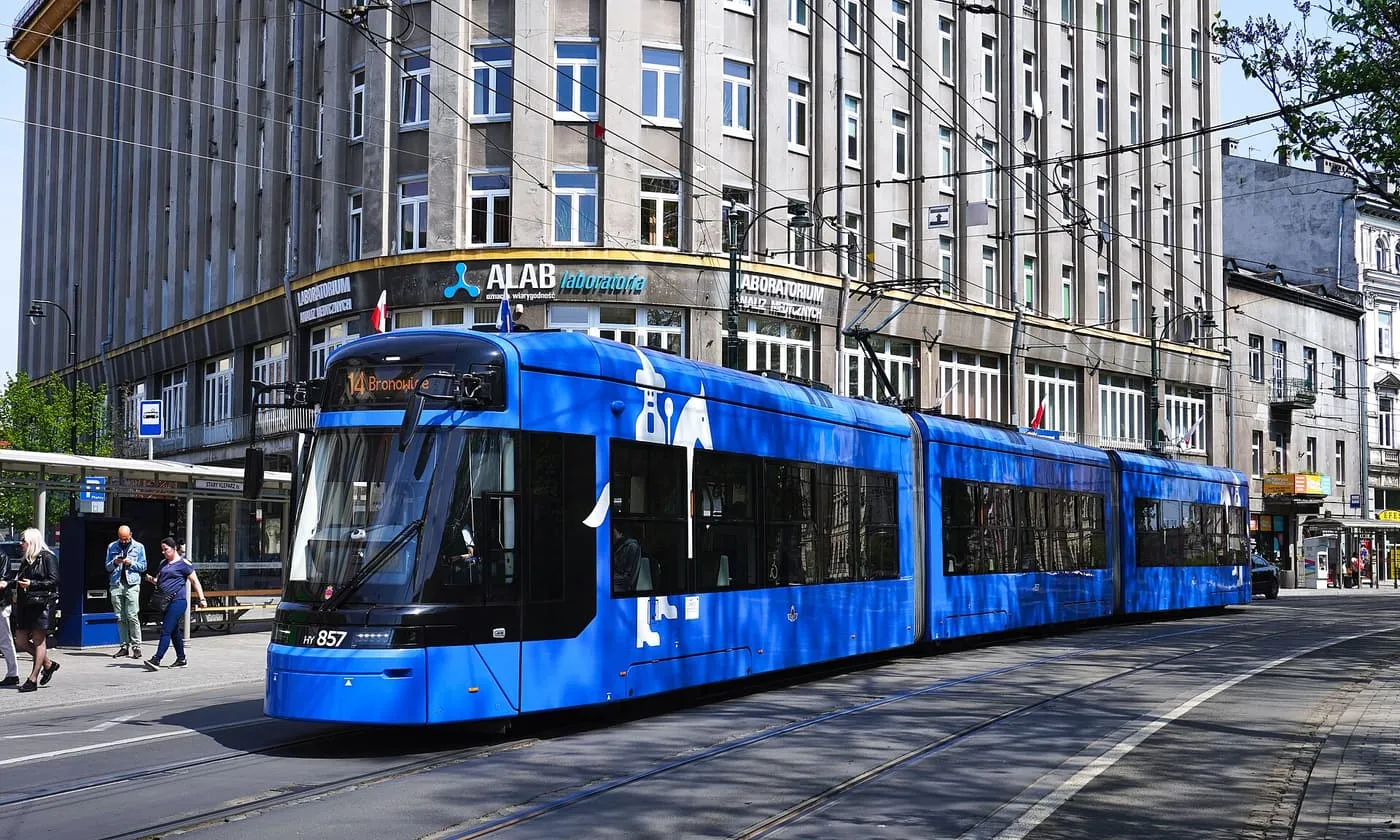 A blue Stadler Tango tram on ul. Basztowa, with a stop and pedestrians beside it and a tall office building behind