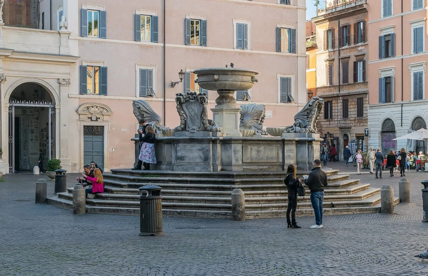 The fountain on Piazza di Santa Maria in Trastevere, with its stepped base, pink and ochre buildings behind and people sitting on the steps