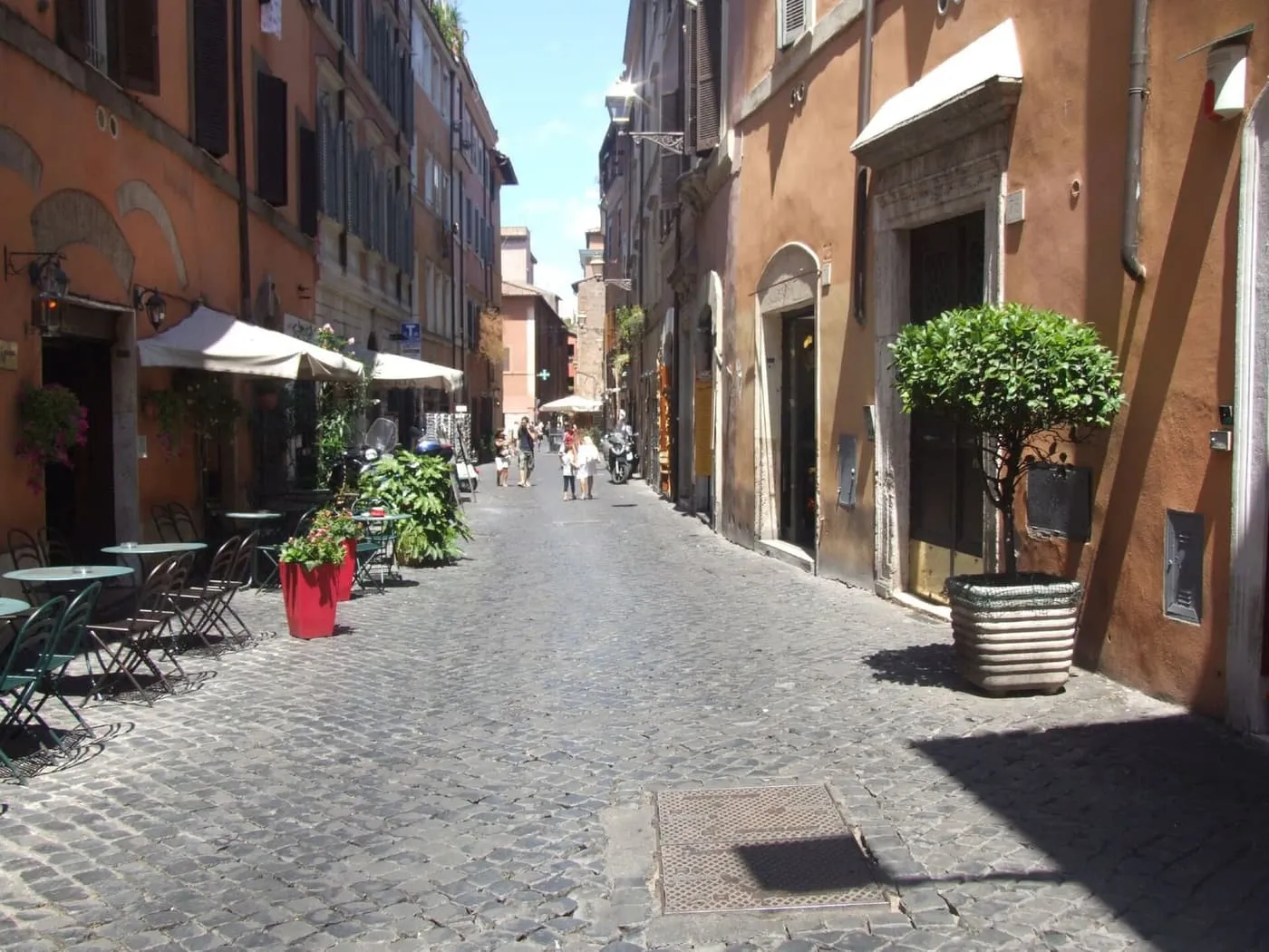 A cobbled street in Trastevere lined with ochre buildings, café tables and potted trees