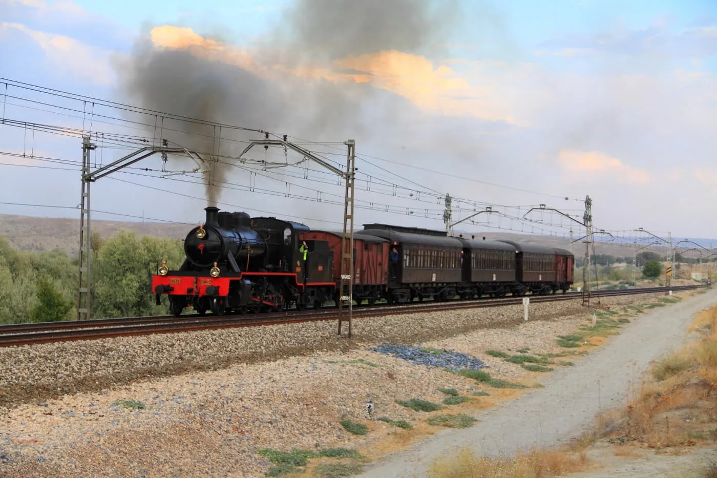 A black steam locomotive with a red front hauling dark wooden carriages along an electrified line, with a plume of smoke overhead