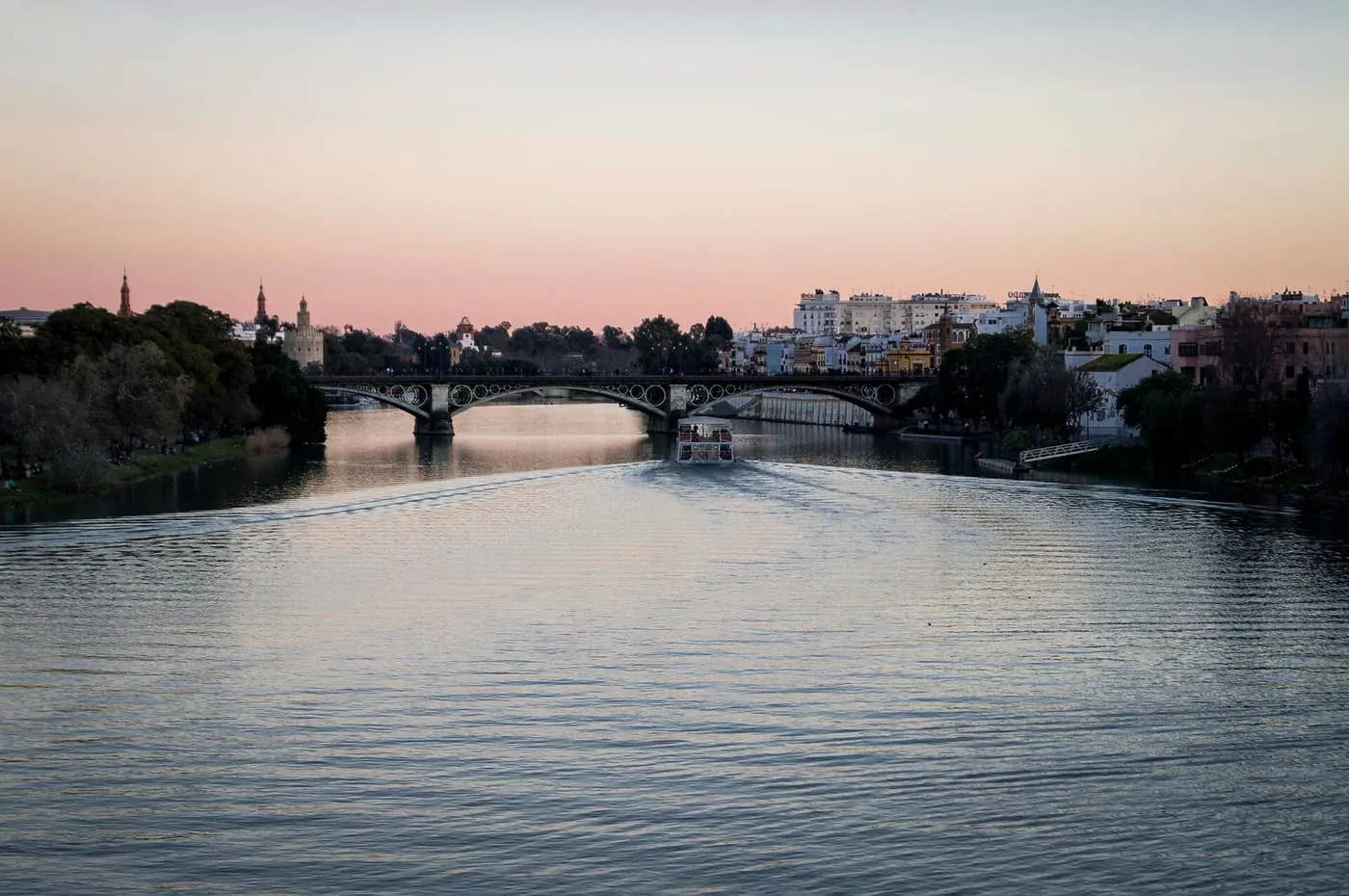 A tourist boat on the Guadalquivir heading towards the Triana bridge at dusk, with the Torre del Oro on the left bank and Triana on the right