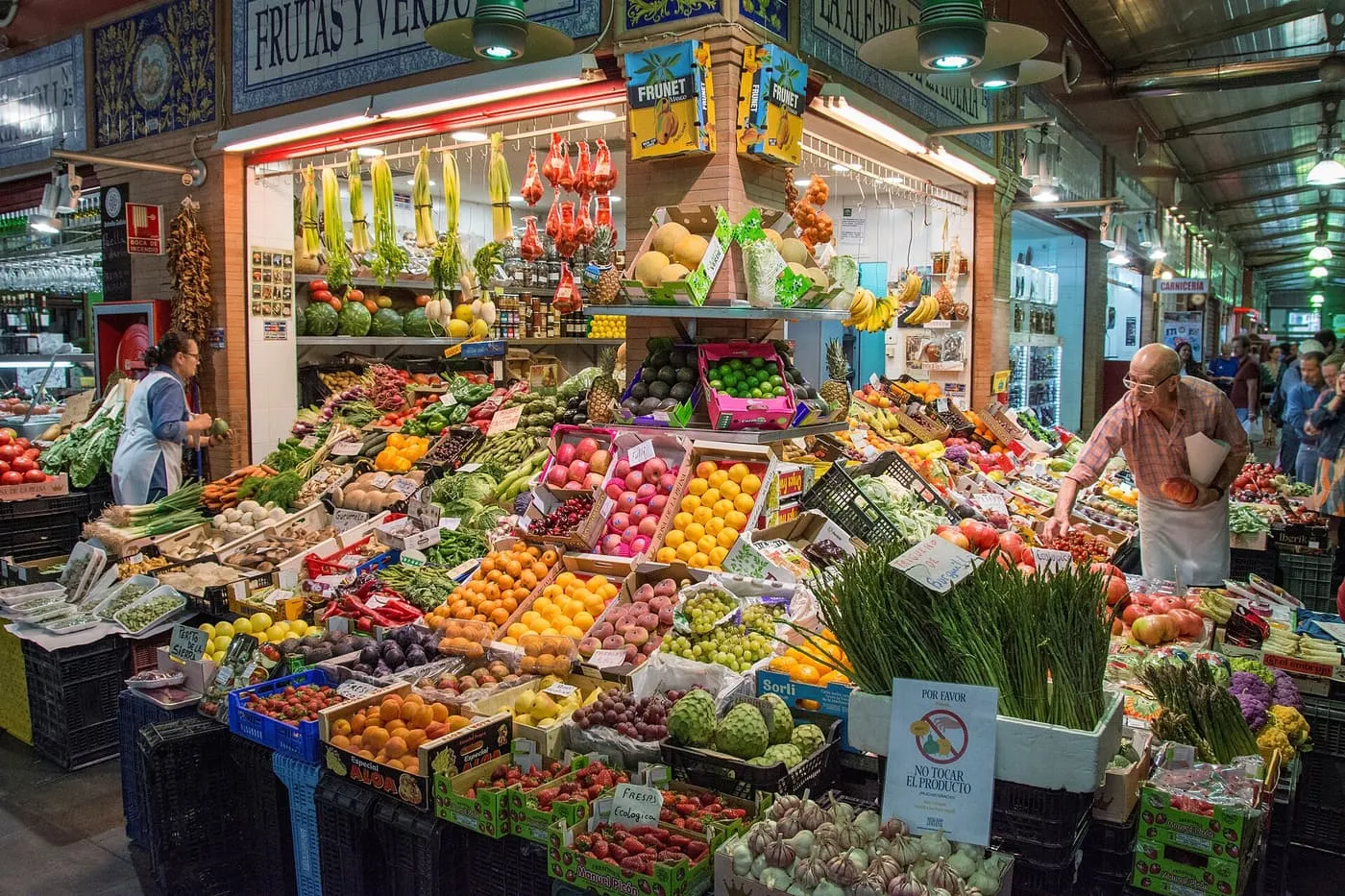 A fruit and vegetable stall in the Triana market piled with peaches, oranges, asparagus and artichokes, with a stallholder at work