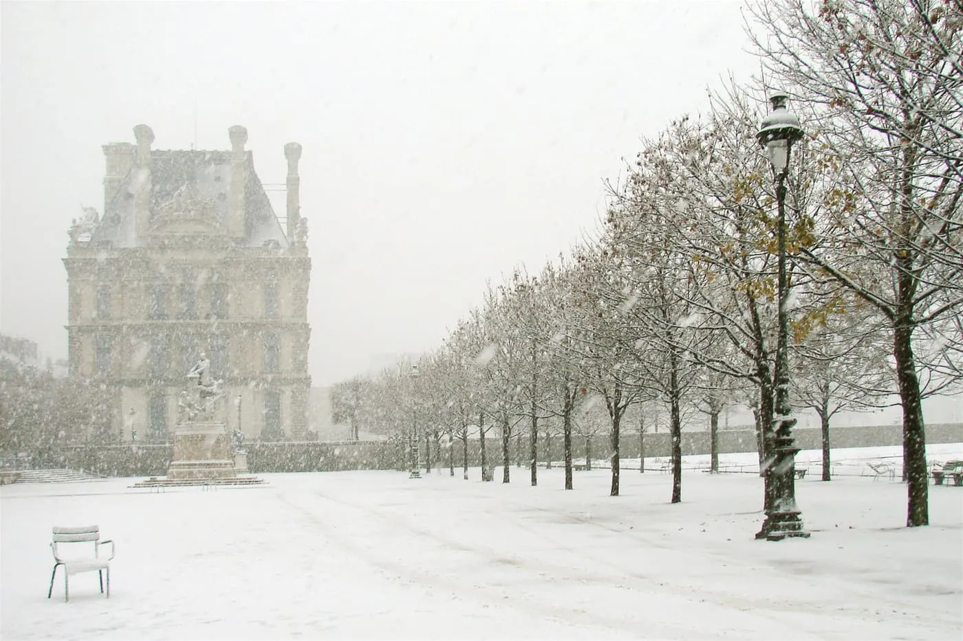 Snow falling on the Tuileries garden, with a snow-covered chair and a row of bare trees beside a lamp post, and a Louvre wing in the haze