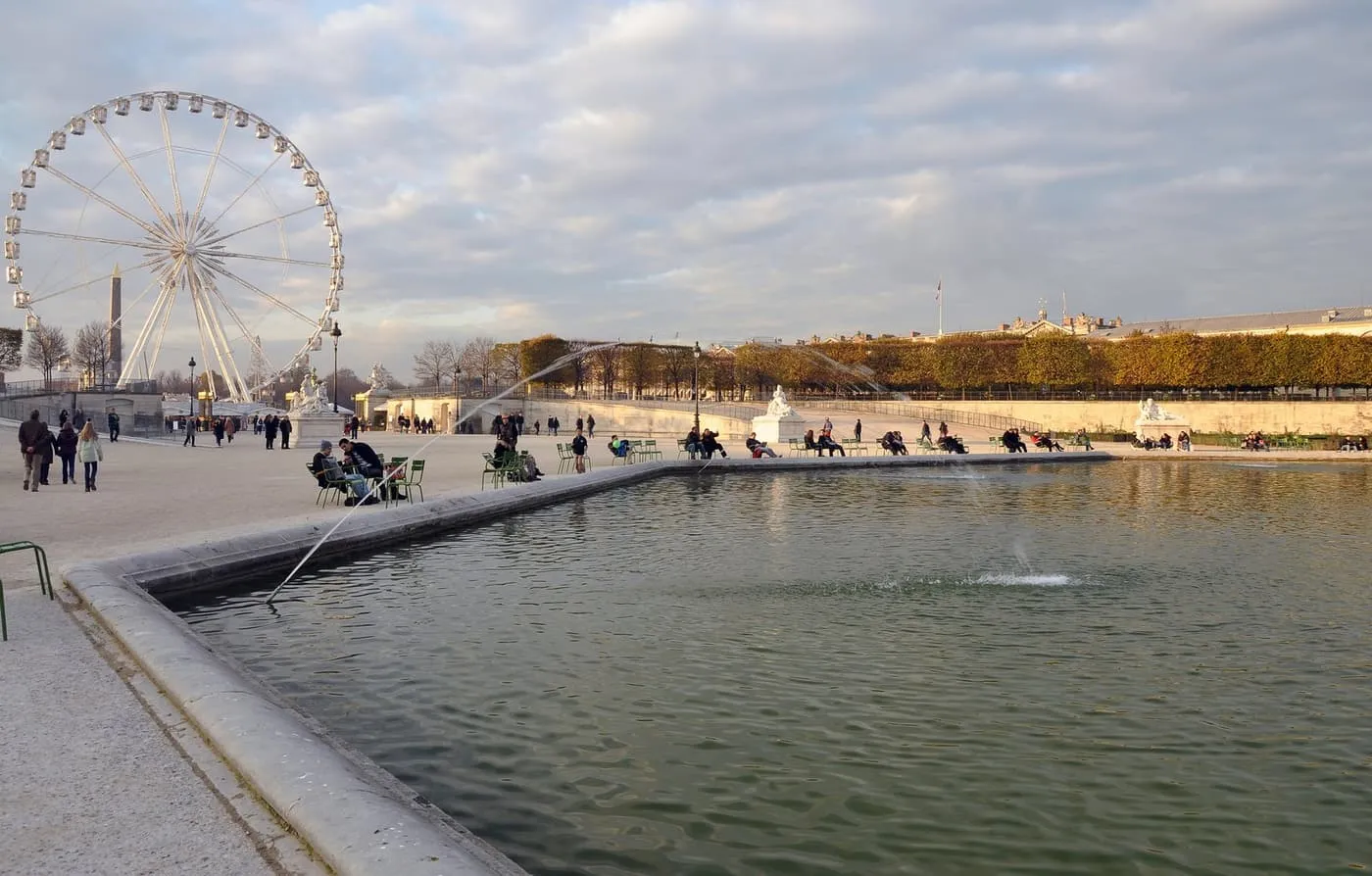 The octagonal pond in the Tuileries garden with a fountain jet, a Ferris wheel and the Luxor Obelisk at the far left, and people sitting by the water