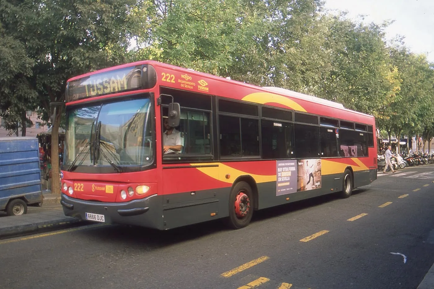 A red and yellow Tussam city bus at a kerb in Seville, with trees behind