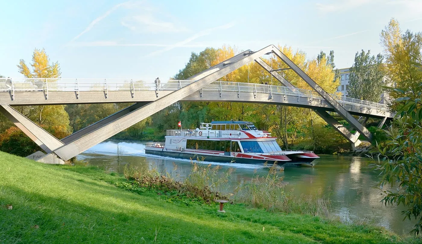 A red and white Twin City Liner catamaran passing under a wooden footbridge between autumn trees